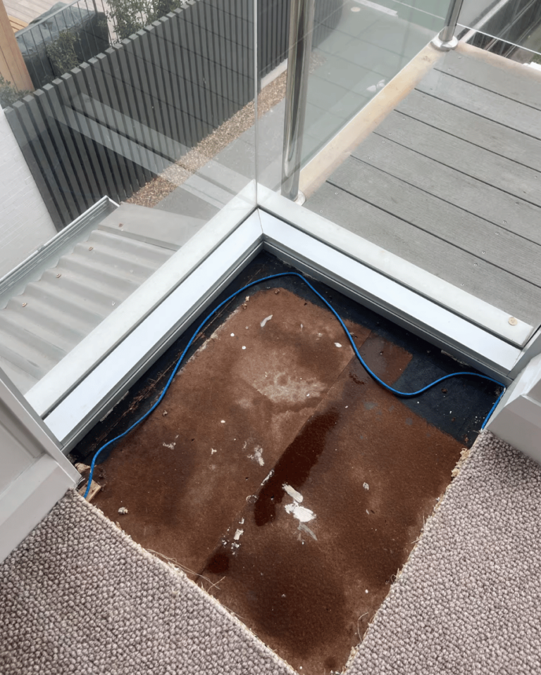 Exposed wet subflooring and stained underlay beneath pulled-back carpet near a leaking glass balcony door tracker during a water damage restoration inspection.