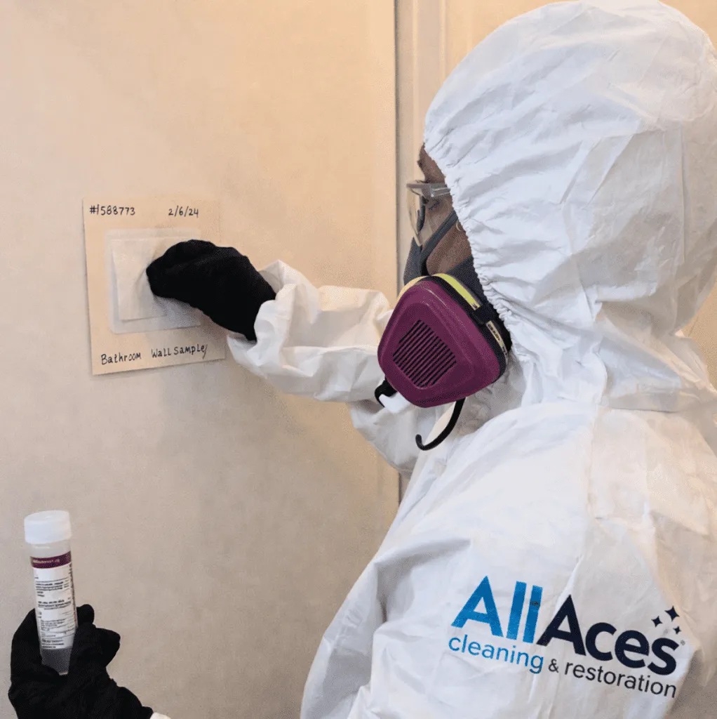 A technician from AllAces Cleaning & Restoration in a white hazmat suit and respirator taking a surface swab sample from a bathroom wall for meth decontamination and testing.